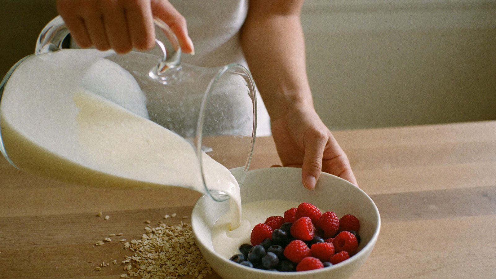 Hands pouring plain yogurt into a bowl with fresh berries and oats